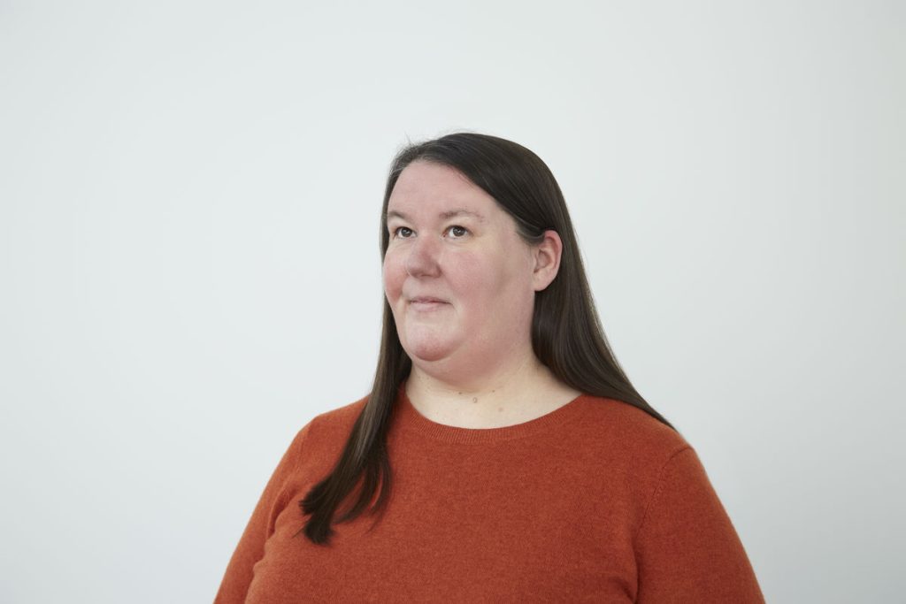 Portrait of Charlotte Cooper by Christa Holka. Fat, middle aged, long dark hair, white, orange jumper, looking upwards