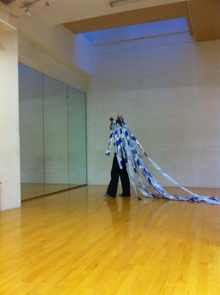 Zinzi Minott in a rehearsal space. She stands on a wooden floor in front of a large mirror. She weaves police tape into her hair.