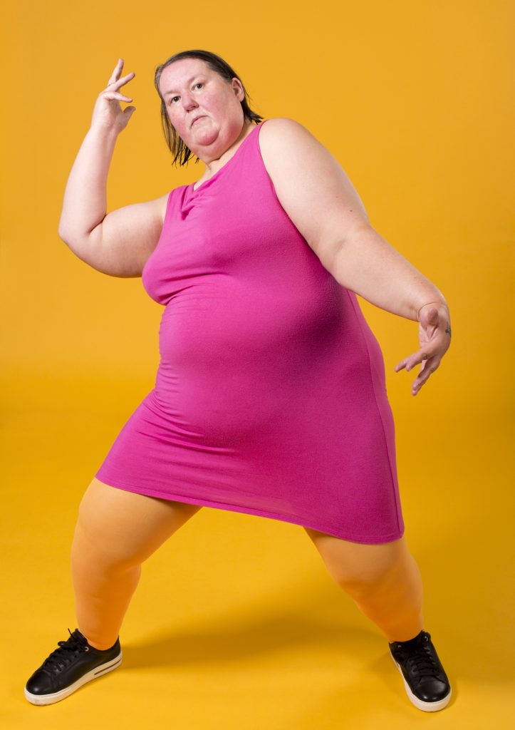 Giant dyke, pink dress, yellow background. Expansive shape, stare, hands.